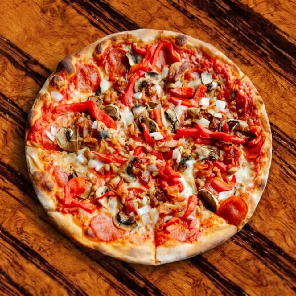 Overhead view of a freshly baked vegetable pizza topped with mushrooms, peppers, onions, tomatoes, and melted cheese on a wooden table.